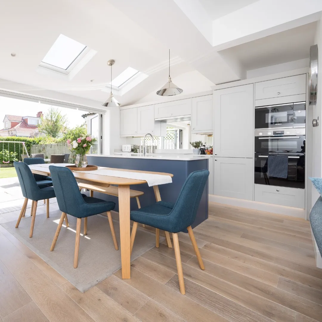 Dining table with blue upholstered chairs beside a navy kitchen island, grey cabinetry with built-in ovens and roof windows above