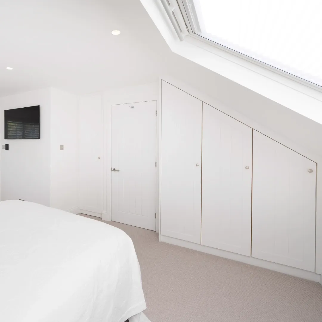 Fitted white cupboards built into the sloping eaves of the loft bedroom beneath a large roof window