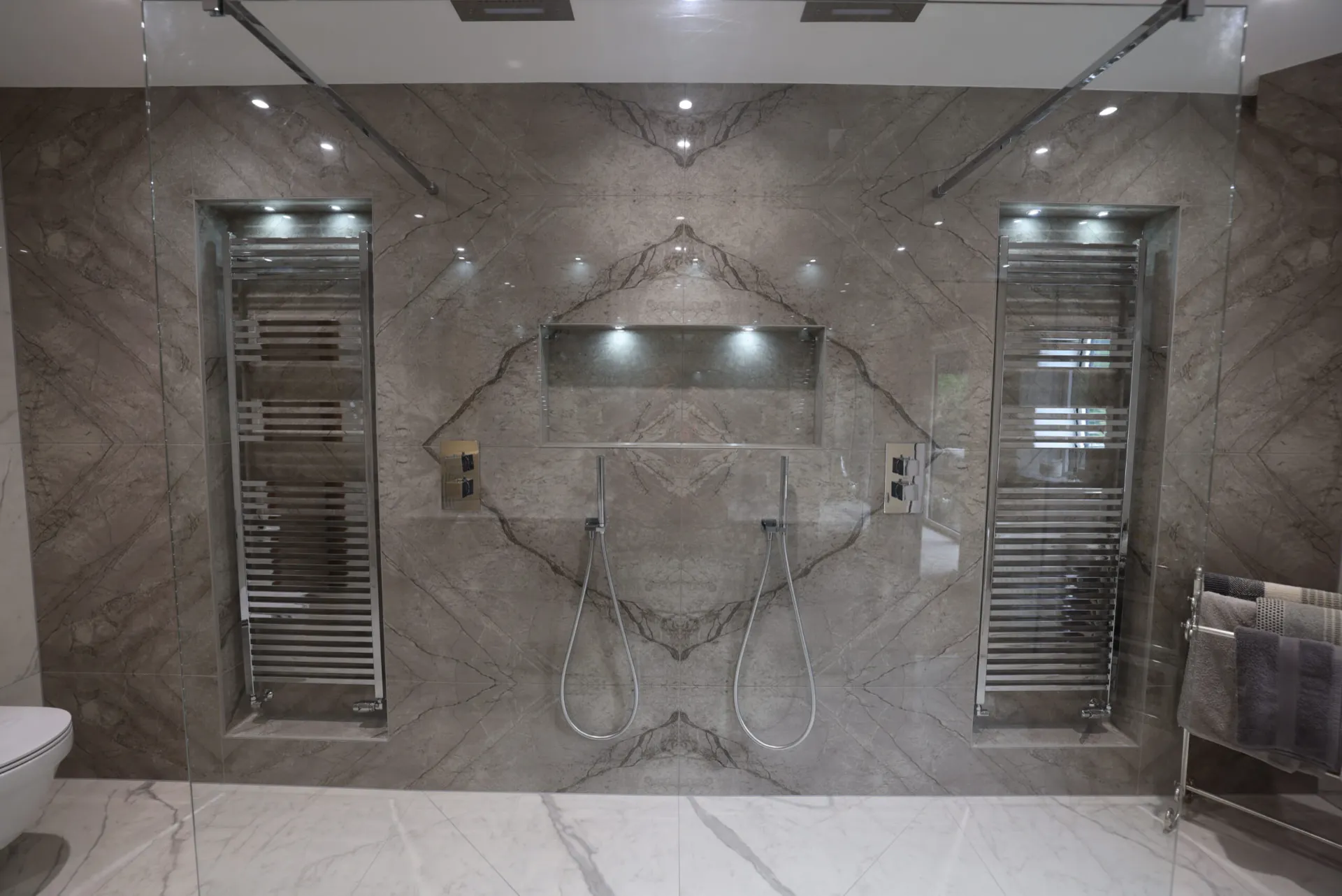 Book-matched stone-effect tiling behind a twin walk-in shower with a lit niche and recessed towel radiators