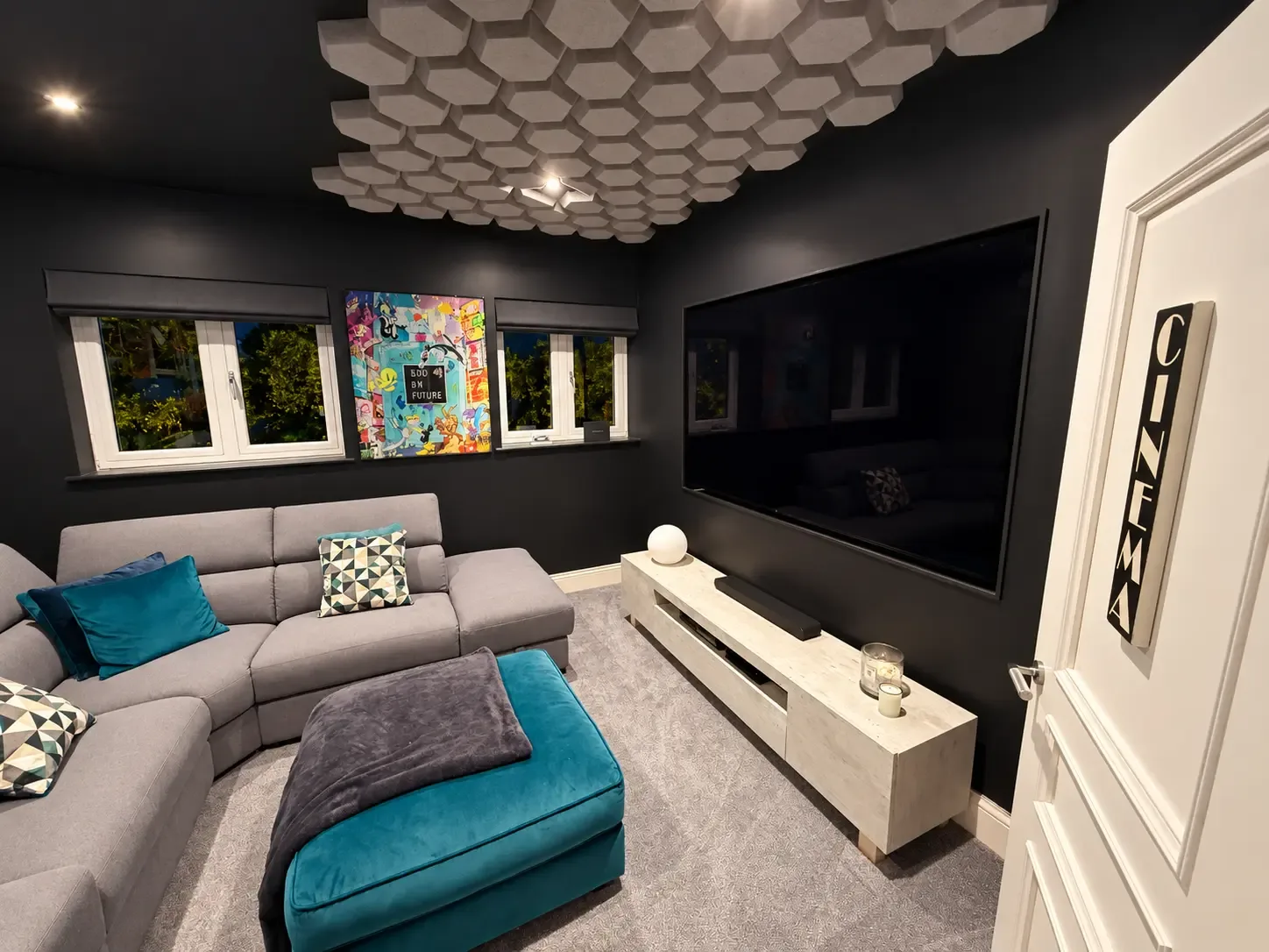 Cinema room with dark walls, a hexagonal acoustic ceiling feature, a large wall-mounted screen, a grey corner sofa and a door marked Cinema