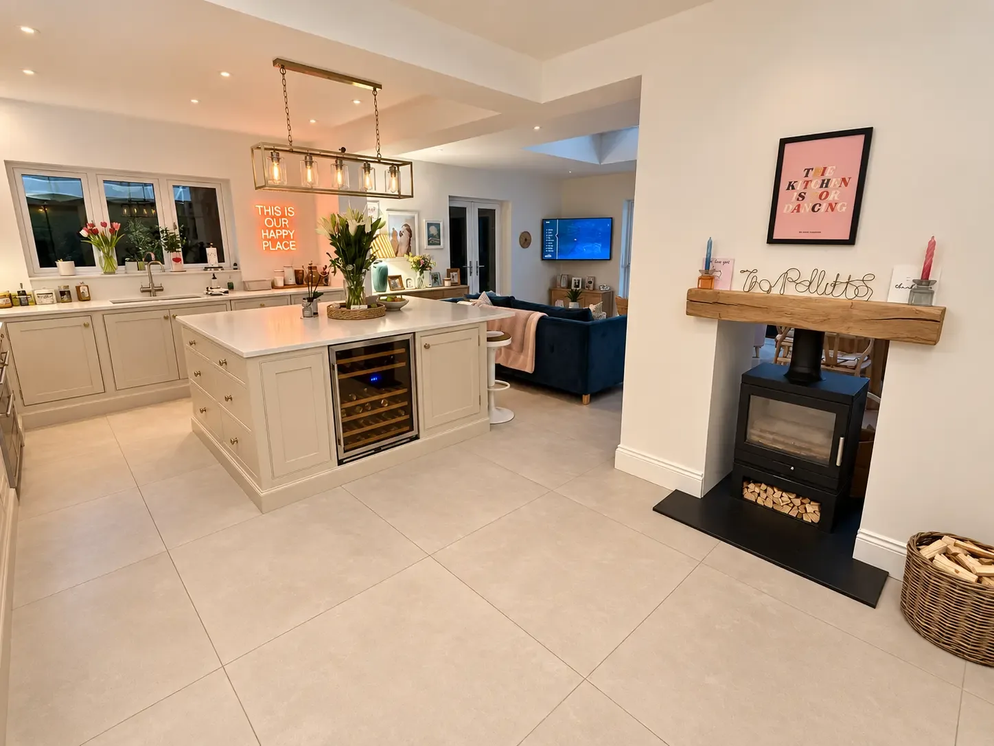 Kitchen island with an integrated wine fridge beside a wood-burning stove with an oak beam mantel, and a seating area with a sofa and wall-mounted television beyond
