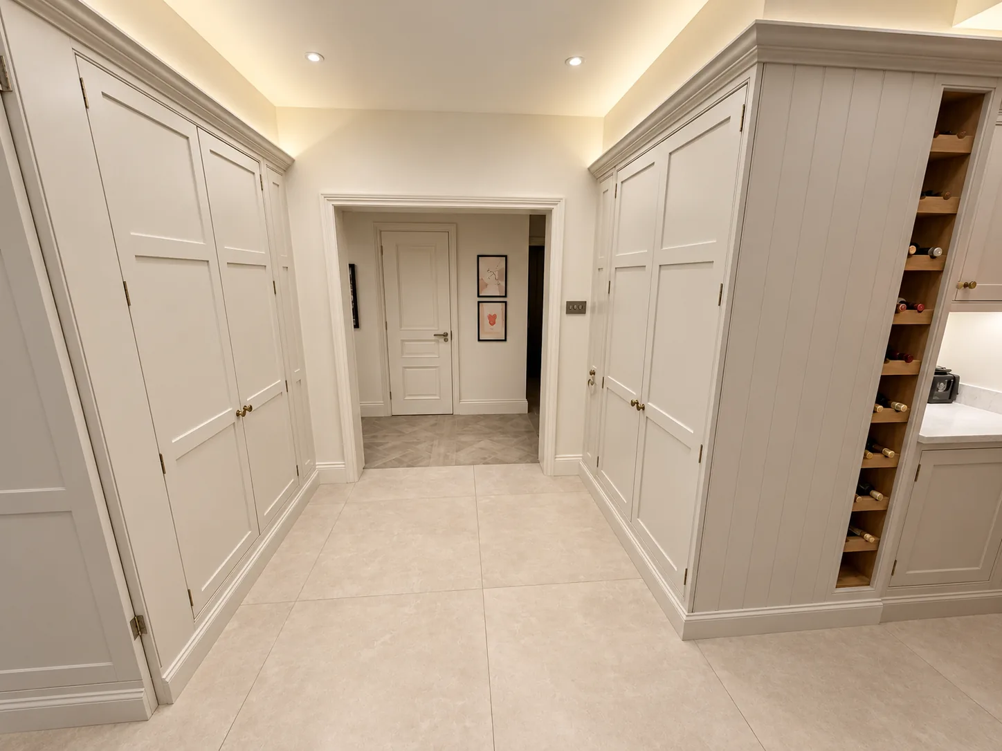 Bespoke full-height painted pantry cabinetry with brass handles and an integrated wine rack, set beneath concealed ceiling lighting with a doorway to the hallway beyond