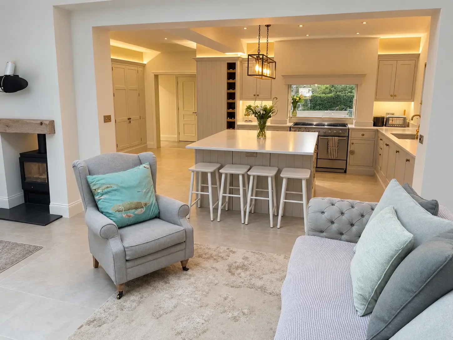 Living area with a grey wingback chair and sofa beside a wood-burning stove, opening through a wide archway into the kitchen and island
